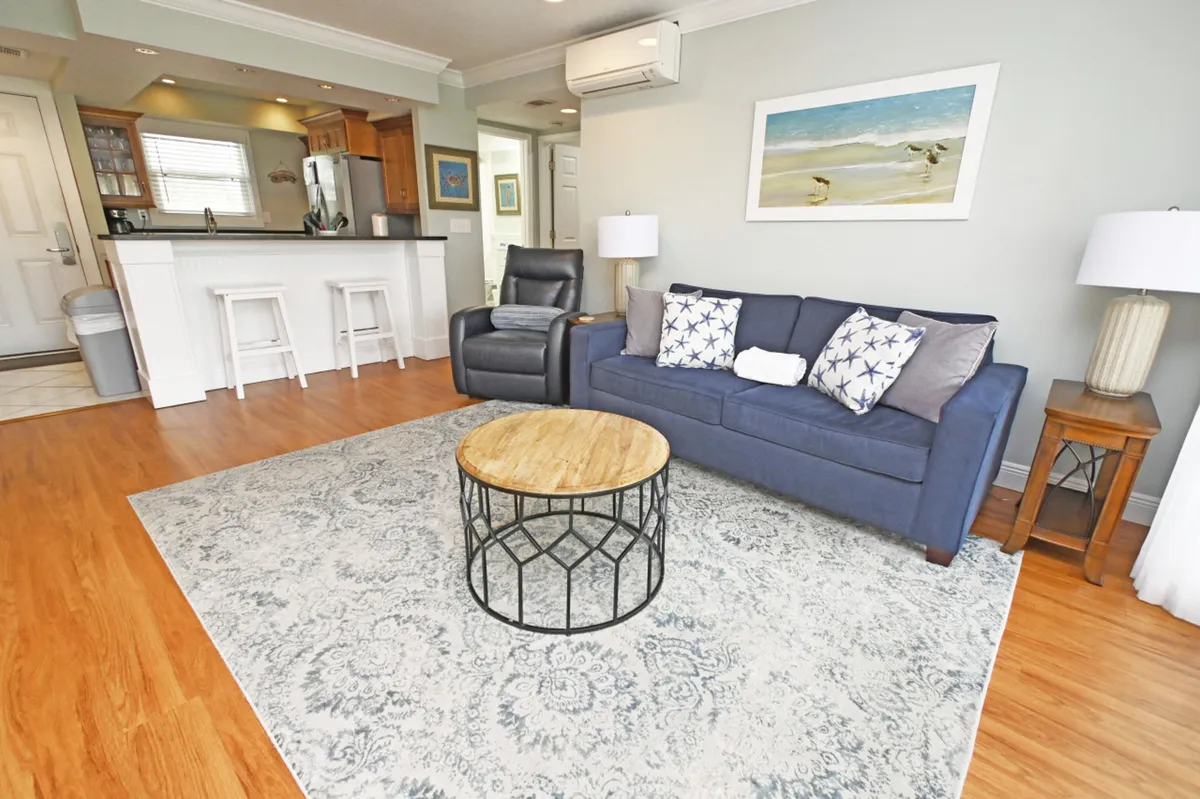 The image showcases a bright, inviting living area featuring a plush blue sofa adorned with starfish-patterned pillows, a stylish round coffee table with a metal base, a cozy recliner, and bar stools at the kitchen counter. The decor blends coastal charm with modern comfort, creating a relaxed, welcoming atmosphere ideal for unwinding after a day at the beach.
