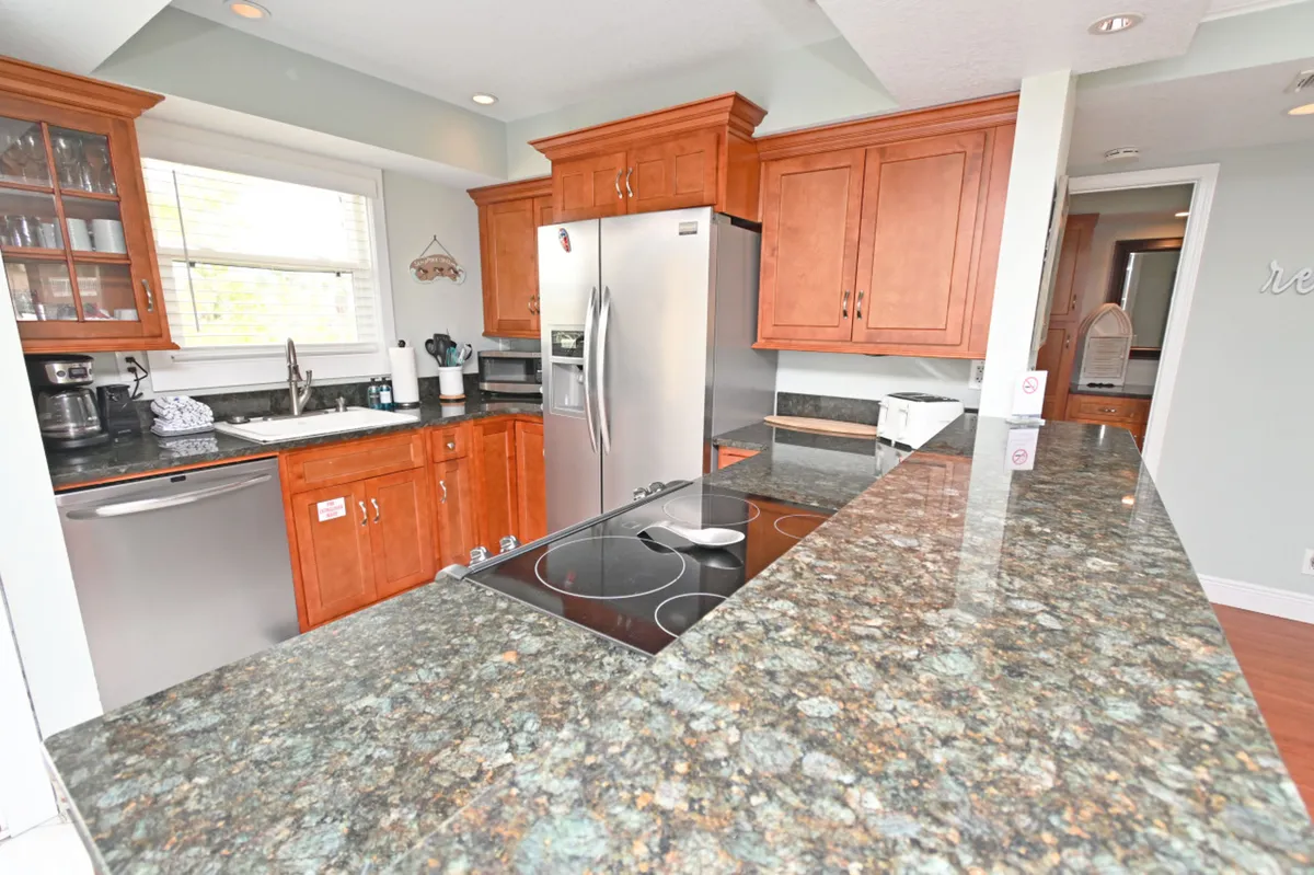 A welcoming kitchen with a granite countertop, built-in electric stove, stainless steel refrigerator, and cherry wood cabinets. The kitchen sink sits beneath a window, inviting natural light and offering garden views. The space is designed for cooking, meal prep, and socializing at the kitchen bar.