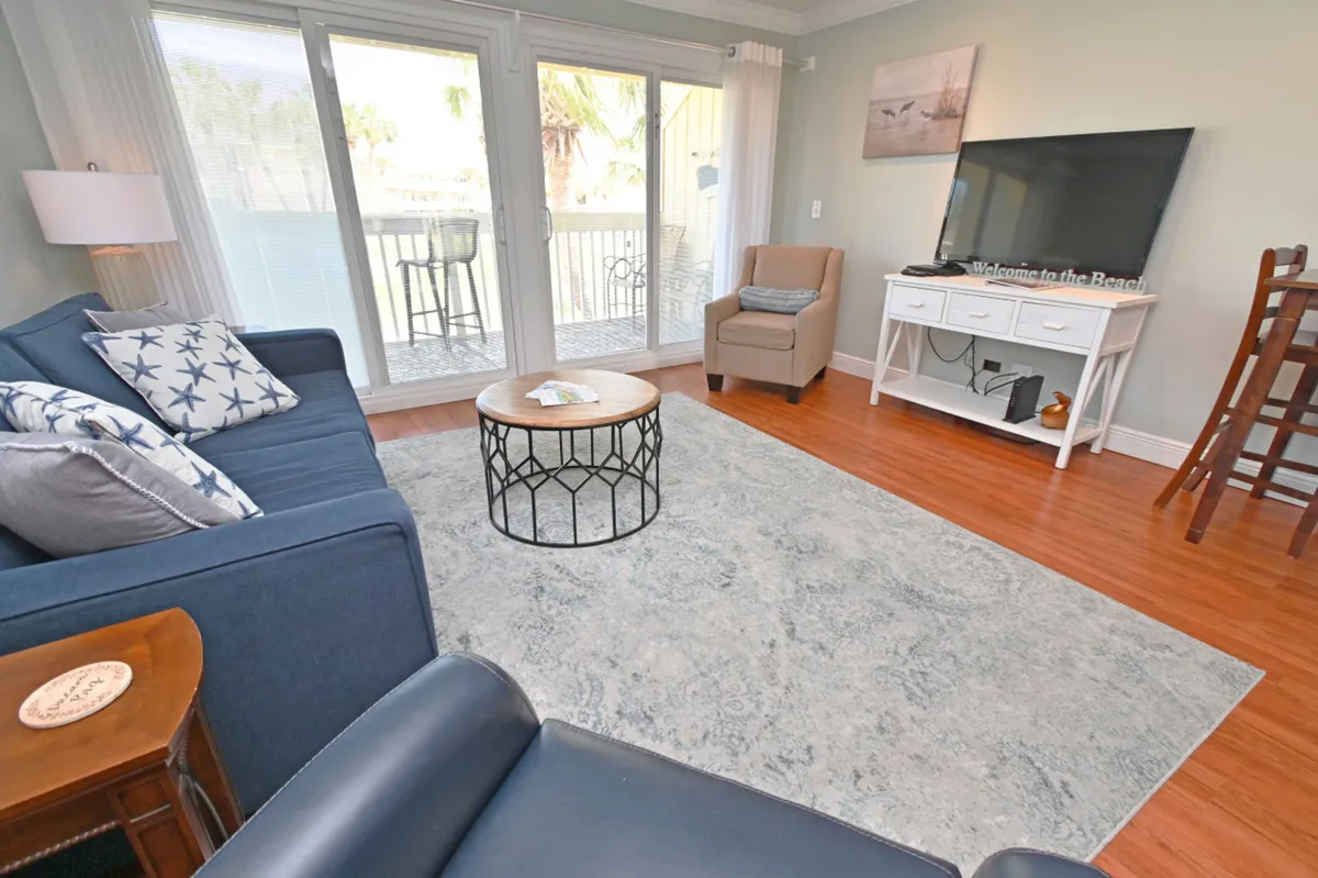 A bright living room with a blue sofa, starfish-patterned pillows, and a round wooden coffee table leads to a balcony through sliding glass doors. The setup invites guests to transition easily between indoor comfort and outdoor enjoyment.