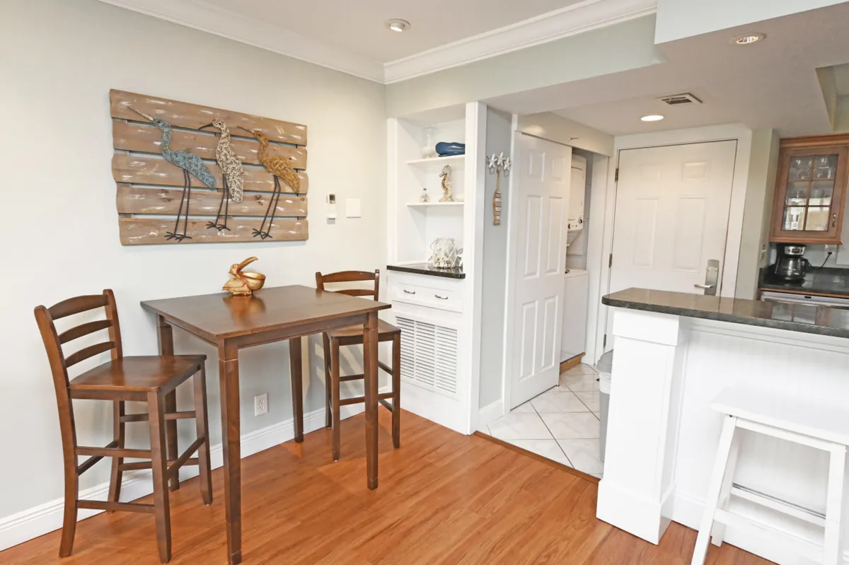 A cozy dining nook featuring a small wooden table with two chairs, coastal-themed wall art, and open shelving displaying decorative items. The kitchen countertop with a bar stool is visible, bridging the dining and kitchen spaces seamlessly.