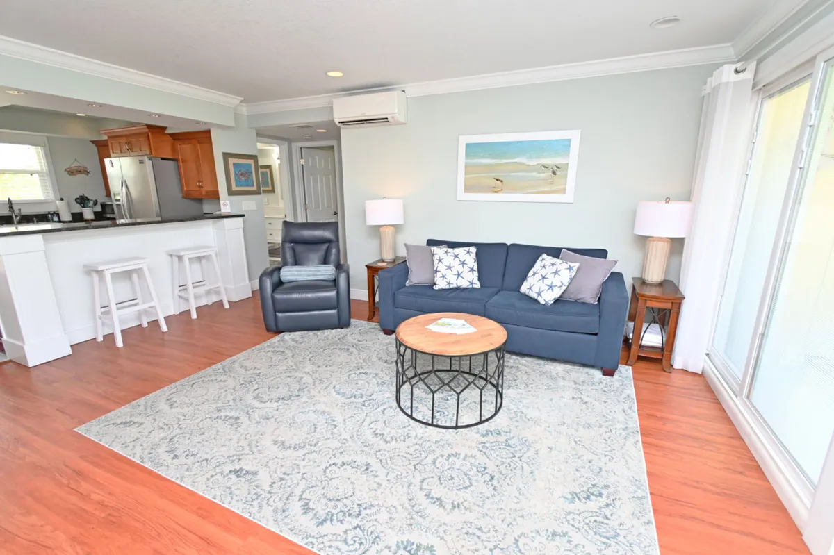 A comfortable living lounge with a navy blue sofa adorned with patterned pillows, a black leather recliner, and a round wooden coffee table. The kitchen bar with white stools and stainless steel appliances is visible in the background, underscoring the open-concept design.