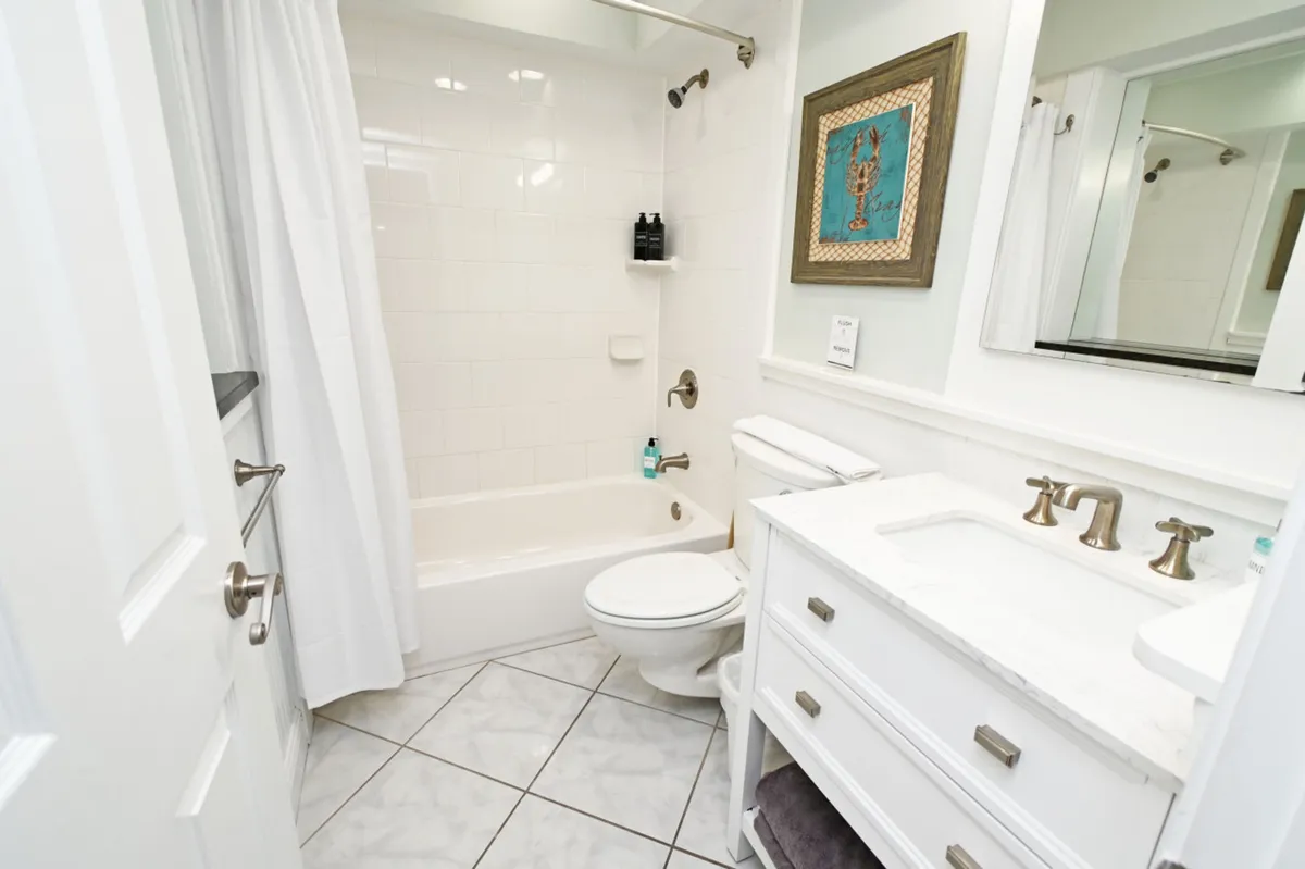 A bright, airy bathroom with a white bathtub and shower, matching toilet, and a white vanity topped with a marble countertop and sink. The space is accented by framed artwork, quality fixtures, and is stocked with plush towels and toiletries for guest comfort.