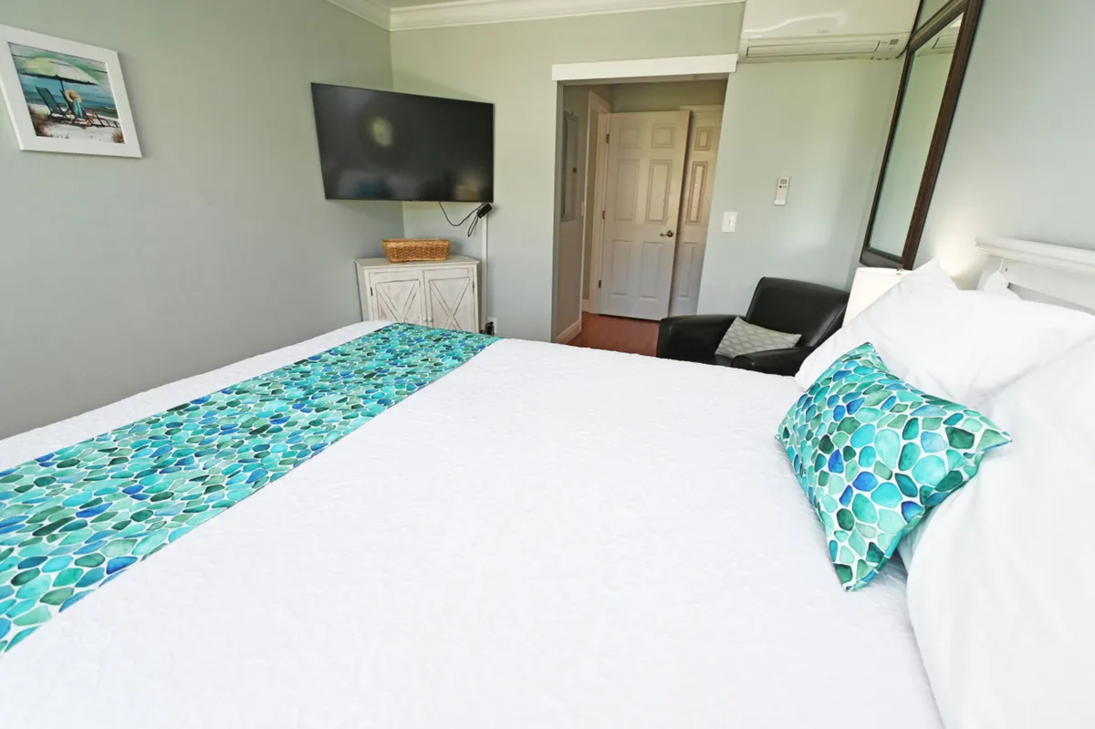 A tranquil master bedroom featuring a queen-size bed with crisp white bedding and teal decorative accents. The room includes a wall-mounted flat-screen TV, a black armchair in the corner for reading or relaxing, and a white dresser beneath the TV, all illuminated by soft, natural light.