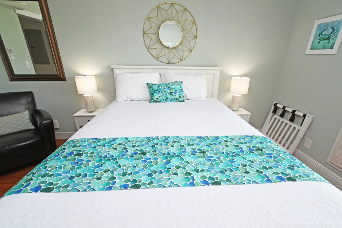 A peaceful bedroom retreat showcasing a queen-size bed with white bedding and a turquoise accent runner. The space is adorned with a decorative sunburst mirror above the bed, two bedside lamps with white shades, and a black leather armchair in the corner, creating a cohesive, calming environment.