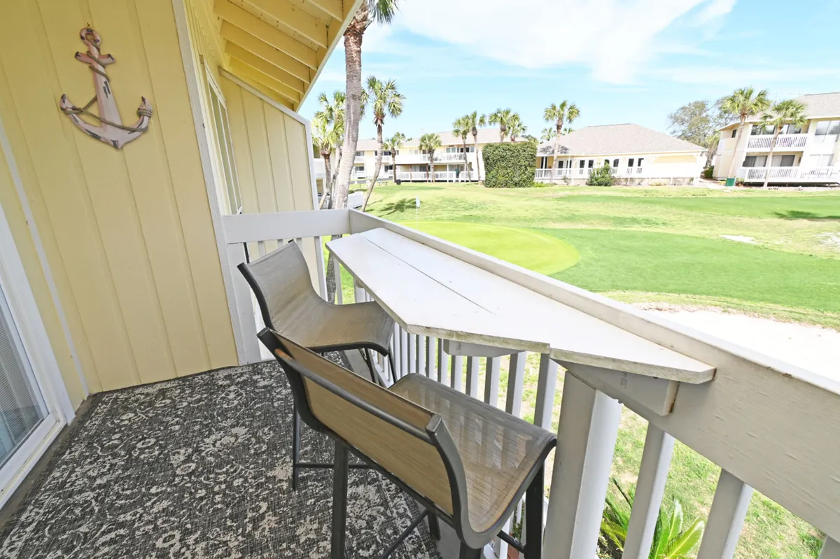 A narrow balcony featuring two chairs and a built-in wooden bar table overlooks the lush green golf course with visible sand traps. The yellow exterior wall is adorned with a decorative anchor, and distant residential buildings and palm trees enhance the scenic view.