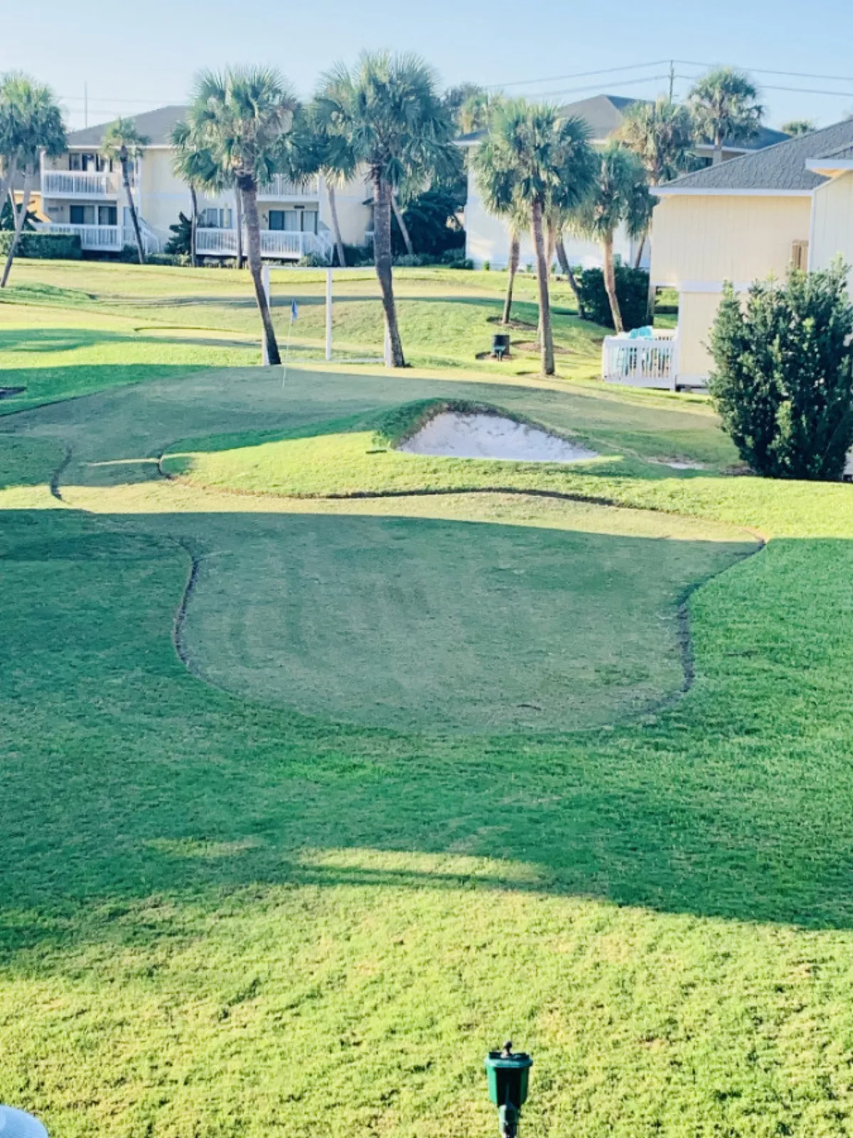 A beautifully landscaped par-3 golf green with a sand bunker, surrounded by palm trees. In the background, residential condo buildings and a clubhouse are visible, highlighting both the recreational and residential elements of the community. The flagstick stands ready for golfers, and the area looks calm and inviting.