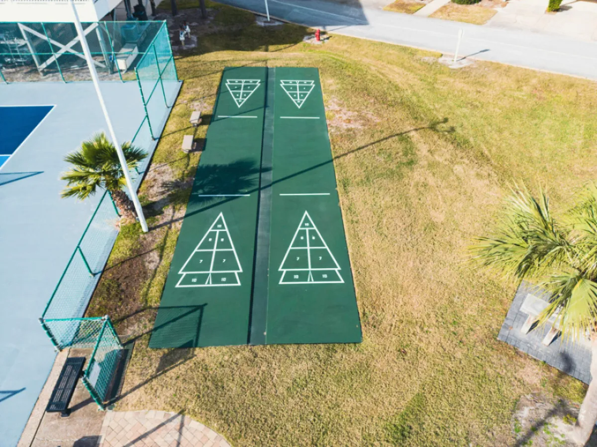 An exterior shot of shuffleboard, tennis, and pickleball courts surrounded by palm trees and benches. The setting encourages active recreation and social engagement, directly referencing the pickleball gear provided within the condo.