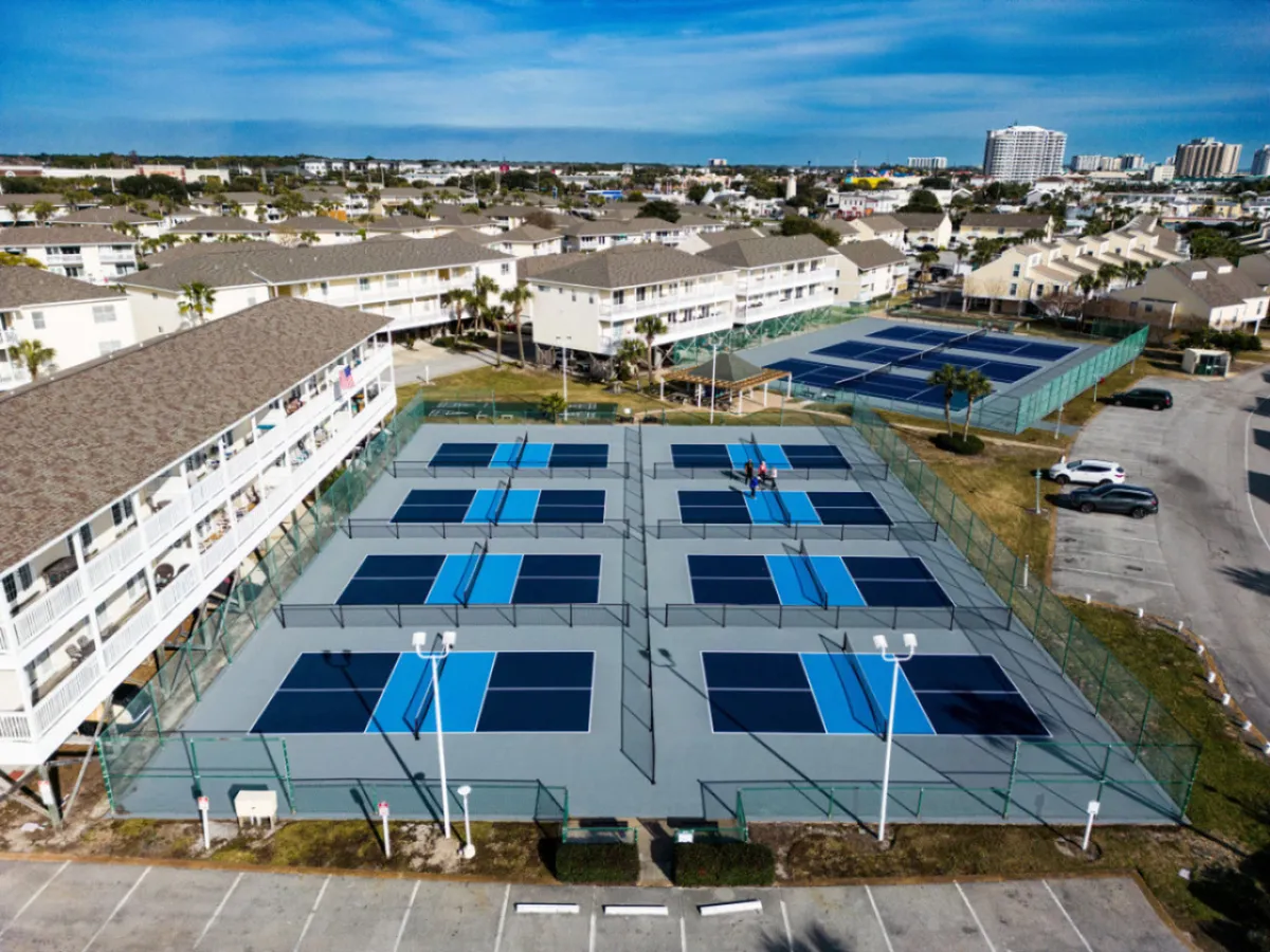 Multiple vibrant blue and navy pickleball courts are surrounded by residential buildings and adjacent tennis courts. The area is active yet orderly, suggesting opportunities for recreation and physical activity. The parking lot and green spaces nearby hint at a convenient and accessible community setting.