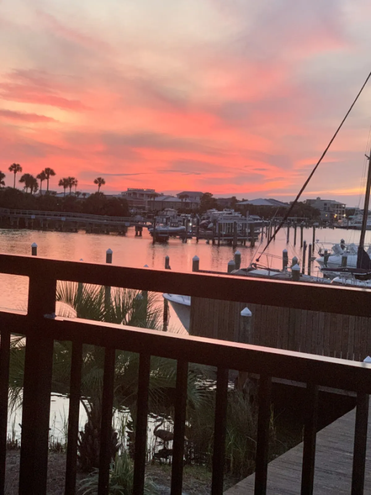 A tranquil sunset scene over the marina, with boats docked at the water’s edge, wooden railings in the foreground, silhouetted palm trees, and a sky awash with vibrant pink and orange hues.