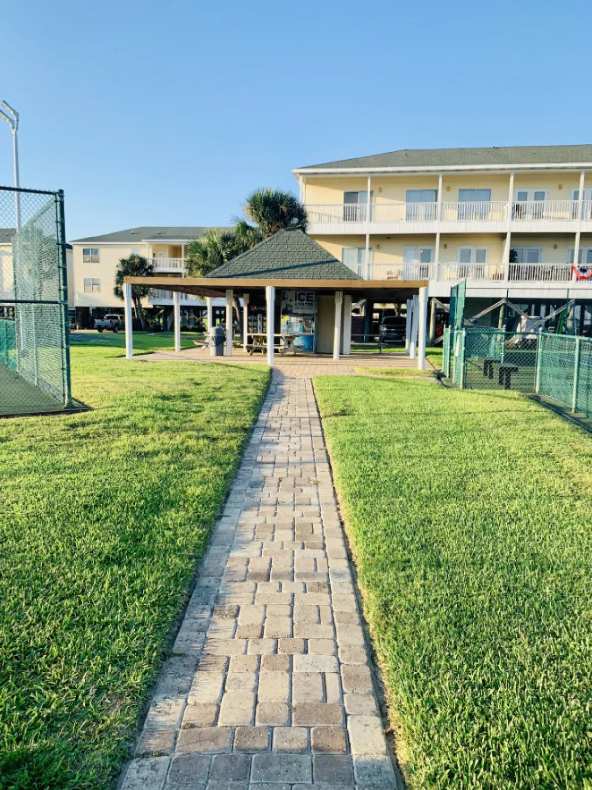 A paved pathway leads to a covered pavilion with picnic tables and vending machines, flanked by fenced tennis or pickleball courts. In the background, a three-story residential building with balconies suggests community living. The courts are ready for play, and the overall scene is relaxed and inviting.