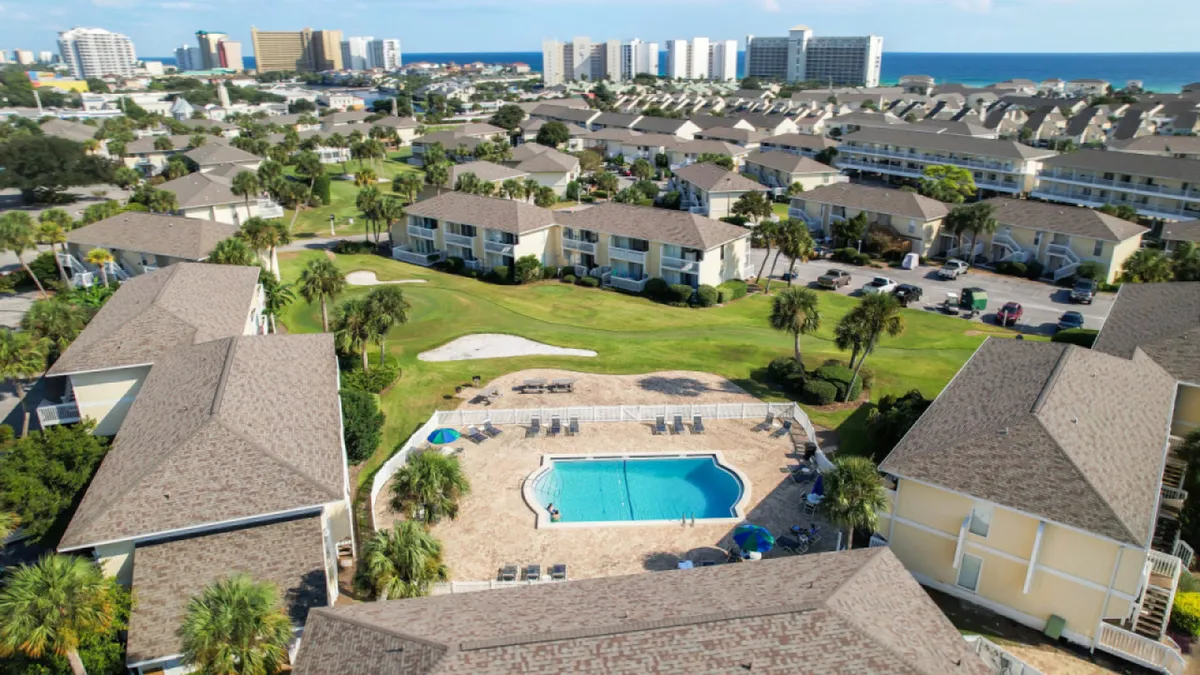 A central swimming pool with lounge chairs is surrounded by well-maintained green space, condominium buildings with balconies, and a visible golf course green complete with a sand trap. In the distance, beachfront high-rise buildings and the ocean are visible, creating a vibrant yet peaceful atmosphere.