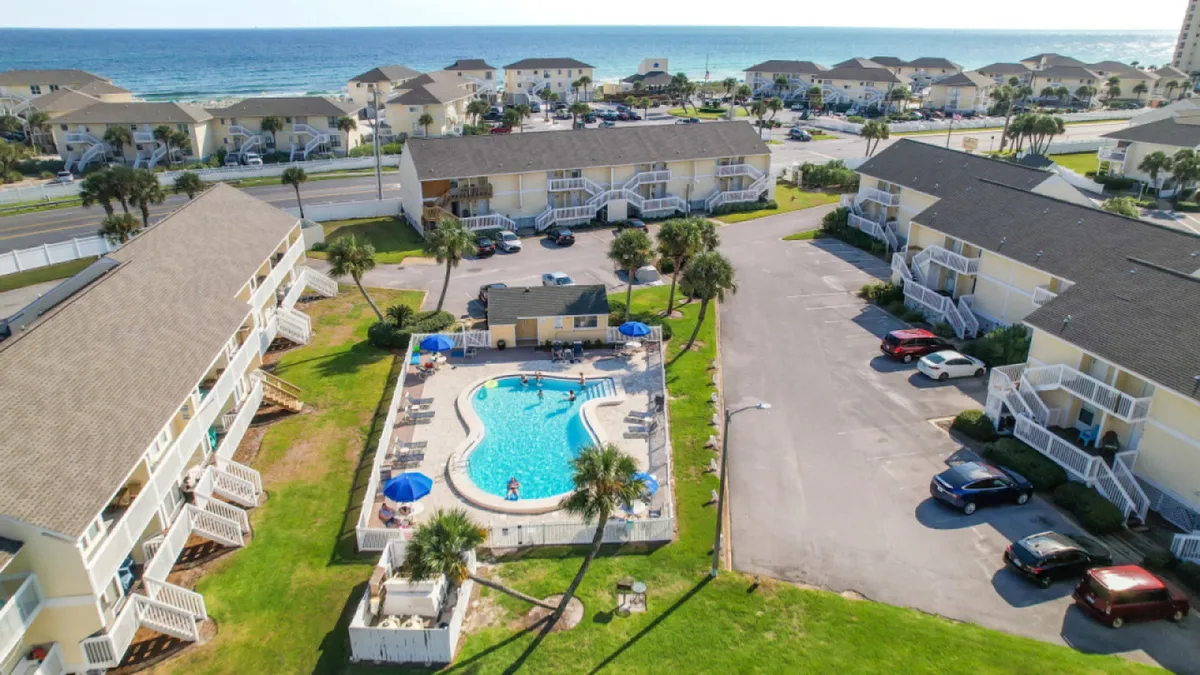 The vibrant scene features a central swimming pool surrounded by lounge chairs and umbrellas, with several low-rise yellow condo buildings, palm trees, and grassy areas nearby. The ocean and a sandy beach are visible in the background, reinforcing the close connection between the pool area and the beach.