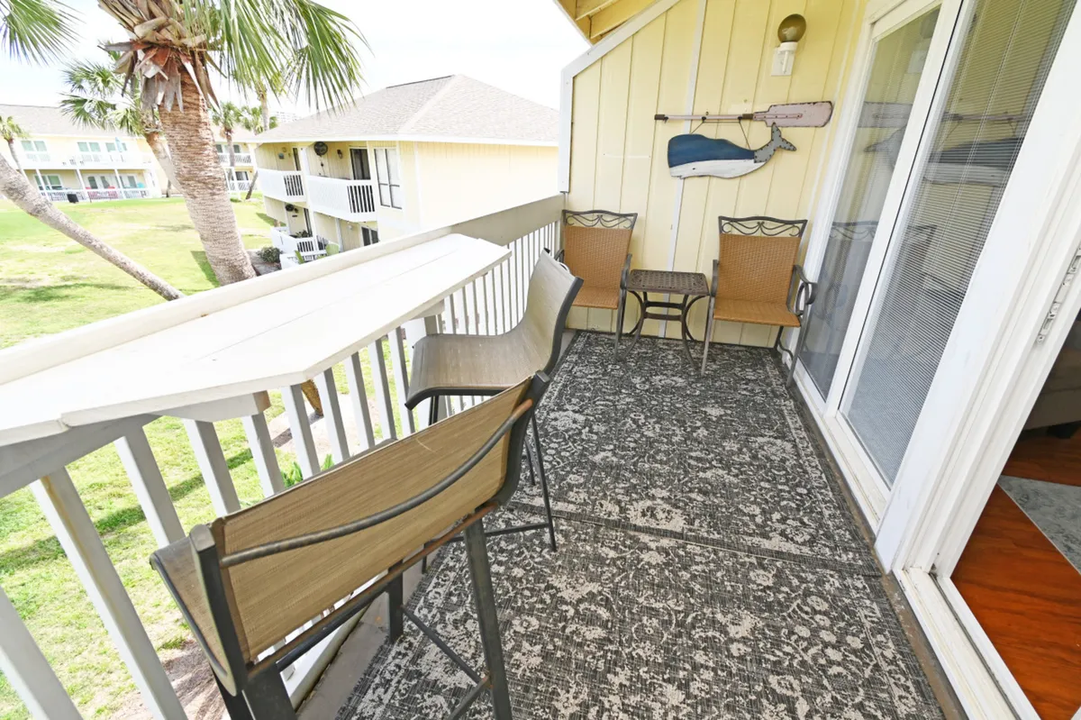 A balcony with bar-style seating for two along the railing and two cushioned chairs with a small table creates an inviting space for outdoor relaxation. Decorative coastal wall art and views of adjacent buildings and palm trees evoke a vacation vibe.