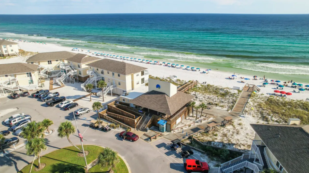 This image features a pristine white sandy beach lined with rows of colorful umbrellas, crystal clear turquoise ocean waves, and multi-unit beachside buildings with balconies. A wooden boardwalk leads directly to the beach, capturing both the ease of access and the visual appeal of beachfront living.