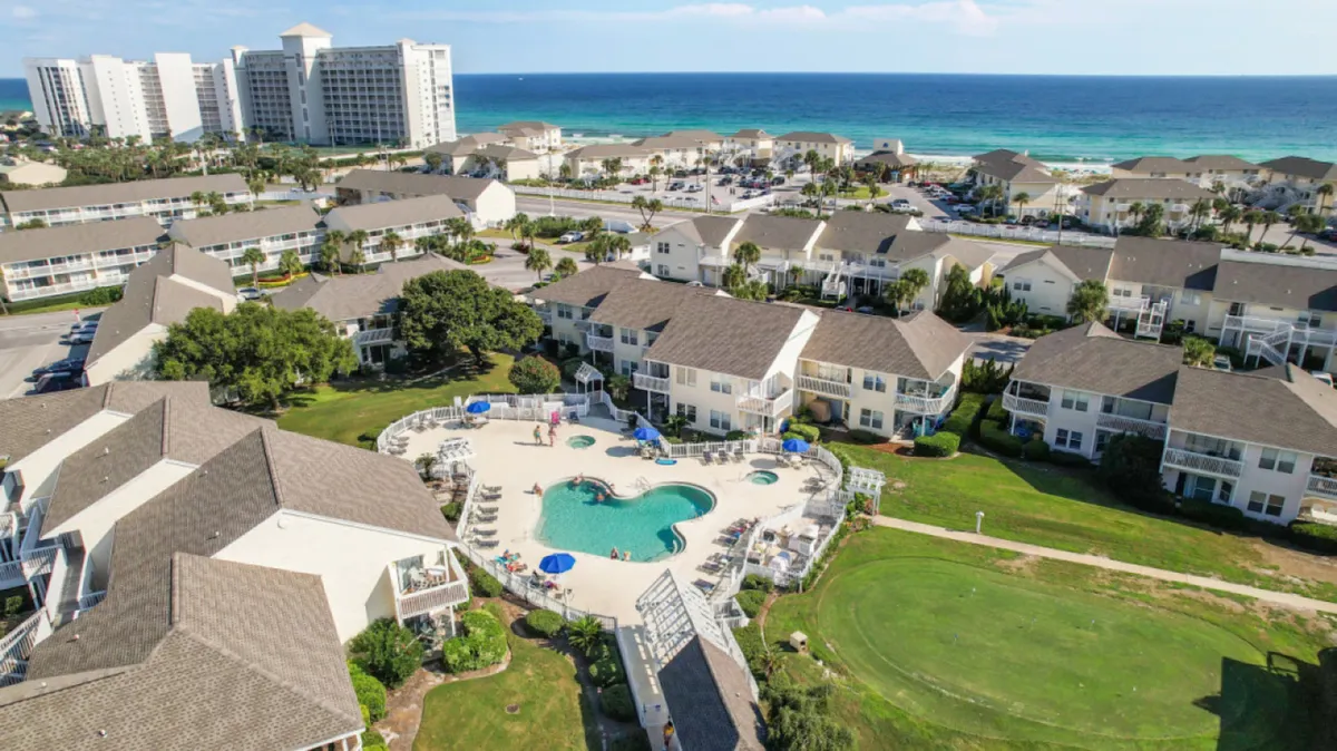 A heart-shaped community swimming pool is surrounded by beige and white condominium buildings, lush green golf course fairways, and the blue ocean and sandy beach in the background. The image captures a broad view of Sandpiper Cove’s amenities, including the pool, golf, and proximity to the beach.