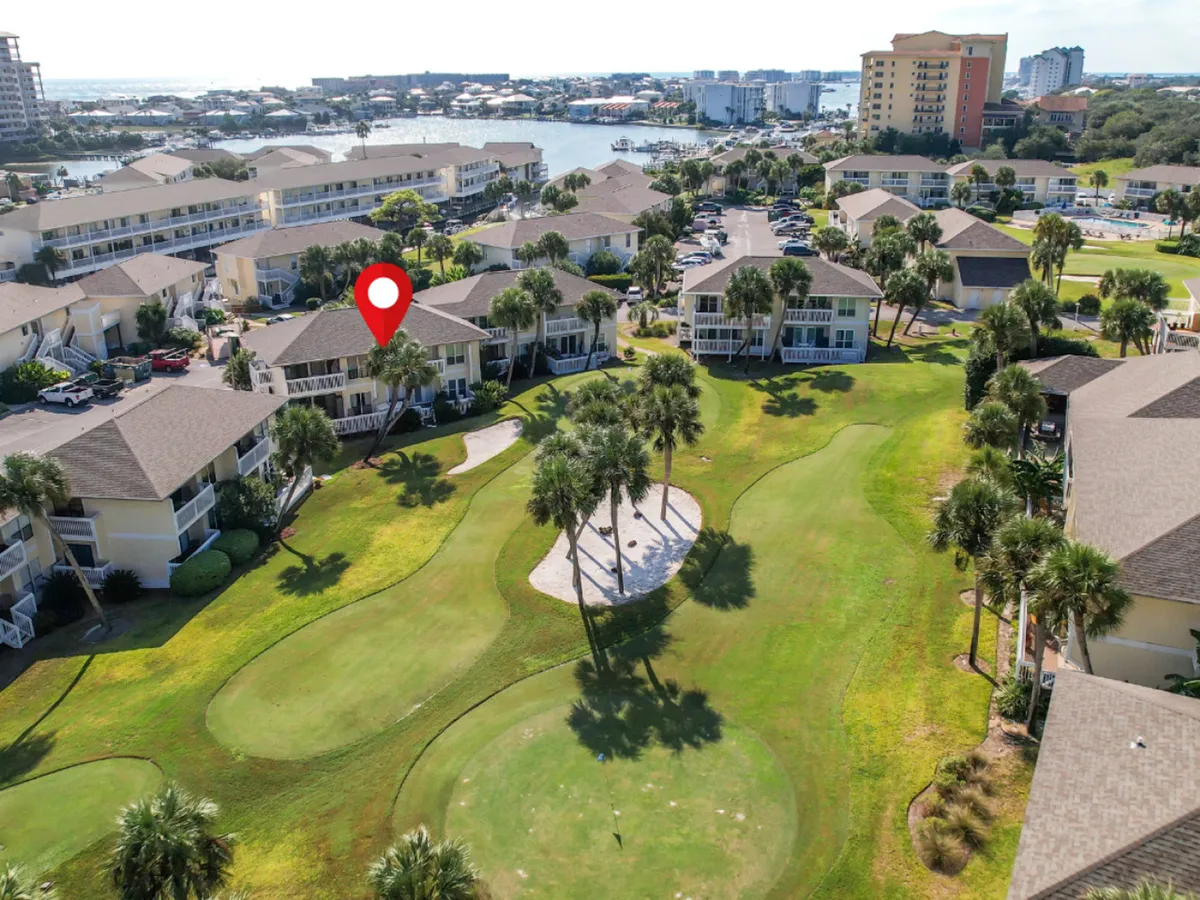 A lush golf course putting green is framed by a cluster of palm trees and nearby condominium balconies, with a marina visible in the distance. The scene suggests leisure and the ability to enjoy tranquil views or a casual round of golf steps from your private retreat.