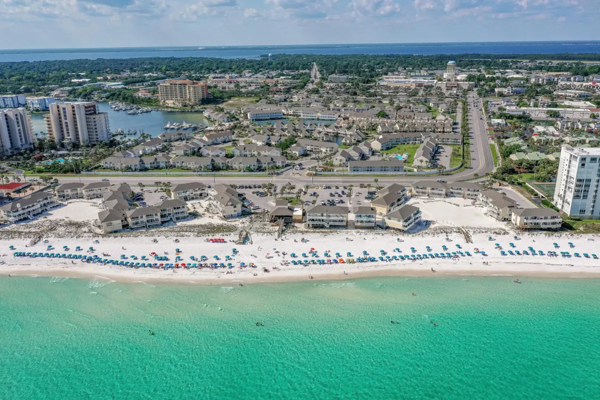 This image presents a lively stretch of white sandy beach lined with colorful umbrellas and chairs, with rows of beachfront condos and townhouses in the background. The turquoise ocean water gently laps the shore under a bright blue sky, and there are groups of people relaxing, swimming, and socializing along the coast.