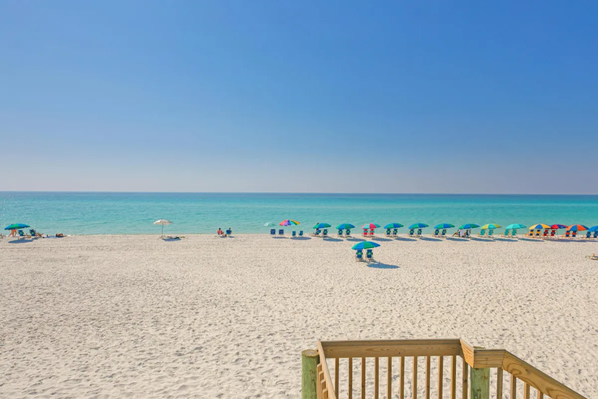 A beautiful stretch of white sand beach lined with rows of vibrant umbrellas and chairs, overlooking calm turquoise waters. The wooden stairs in the foreground indicate easy access from the condo to the beach and its amenities.