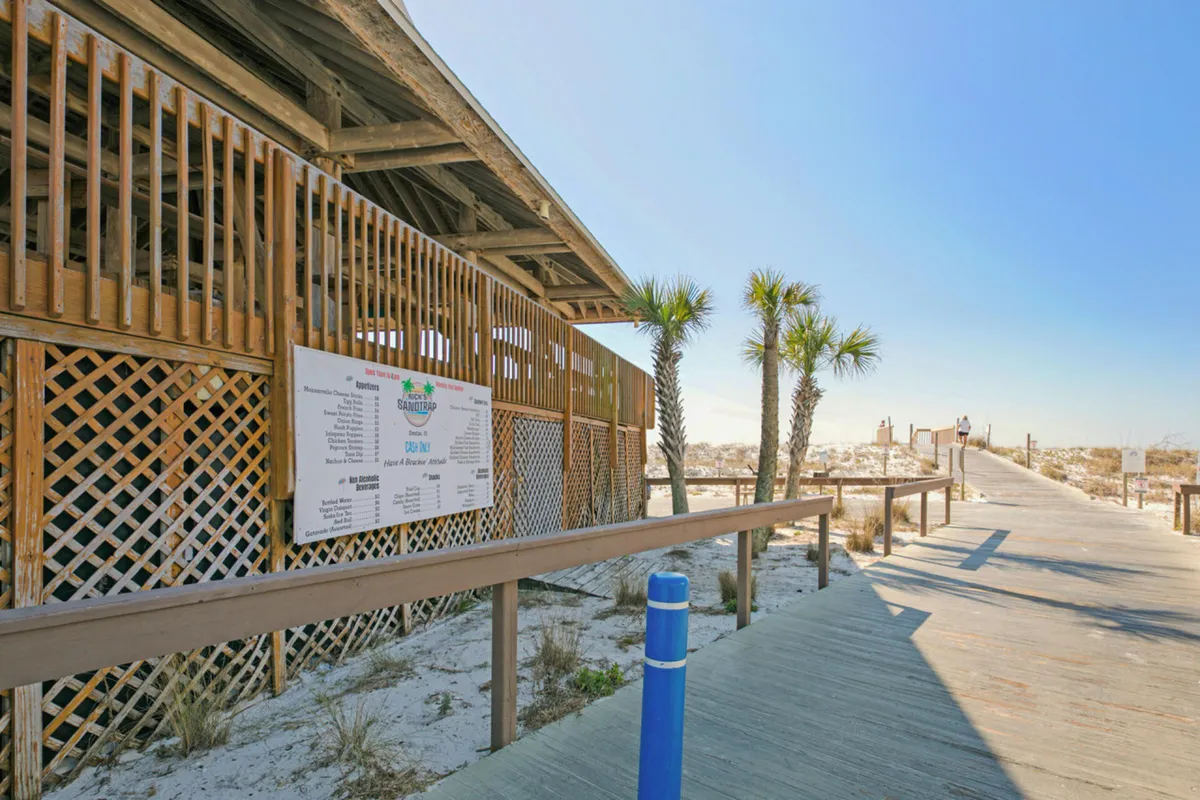 A wooden boardwalk leads toward the beach, flanked by sandy dunes with sparse vegetation and dotted with palm trees. The beach access building, featuring prominent signage, hints at both proximity and convenience for guests. The scene radiates a calm and peaceful mood, inviting visitors to start their day with a scenic walk to the shore.
